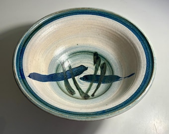 May include: A round ceramic bowl with a cream-coloured interior and exterior. The rim features a thick blue band, while the centre of the bowl is decorated with hand-painted blue and dark green brushstrokes in a simple, abstract pattern.