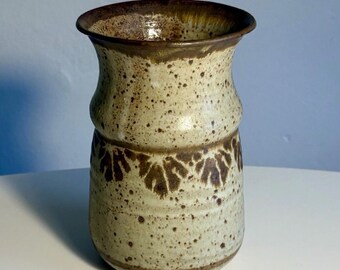 May include: A ceramic vase with a flared rim, featuring a speckled cream and beige glaze. The body is decorated with a horizontal band of dark brown leaf-like patterns, with additional brown speckling throughout the exterior and inner rim.