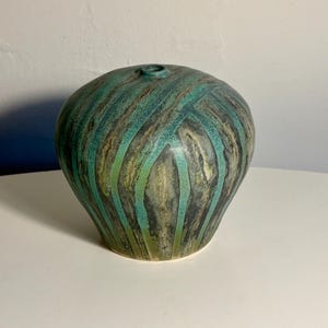 May include: A rounded ceramic vase with a narrow opening at the top. The surface features a textured pattern of vertical stripes in shades of teal, dark green, and earthy brown, creating a layered appearance across the curved body of the vessel.