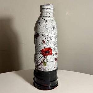 以下が含まれることがあります： 黒い斑点と手描きの赤いポピーの花が描かれた、質感のある白い表面を持つ背の高い円筒形の陶器製ボトル。ボトルの底は光沢のある黒い釉薬で仕上げられており、首の部分には凹凸のある縁があります。