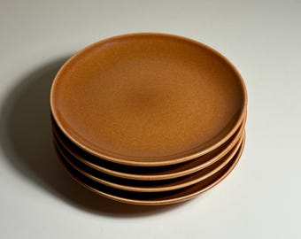 May include: A stack of four round ceramic plates with a speckled, matte, terracotta-coloured finish. Each plate features a slightly raised rim and a smooth, uniform surface texture, creating a consistent appearance throughout the set.