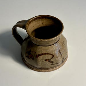 May include: A stoneware mug with a speckled tan and brown glaze. The mug features a wide, faceted body, a sturdy handle, and dark brown hand-painted abstract line markings on the exterior surface. The interior is smooth and dark.