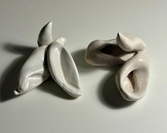 May include: Two white ceramic sculptures with smooth, glossy surfaces. Each piece features organic, curved, and folded shapes that resemble abstract forms or stylised ears. The objects are solid white with subtle shadows.