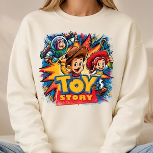 May include: A cream-colored crewneck sweatshirt featuring a vibrant graphic print of Buzz Lightyear, Woody, and Jessie from Toy Story. The design includes a colorful starburst background and the bold blue and yellow Toy Story logo text.