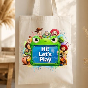 May include: A cream-colored canvas tote bag featuring a colorful graphic of Toy Story characters surrounding a bright green frog-shaped frame. The center of the frame contains the blue text "Hi! Let's Play" against a light blue background.