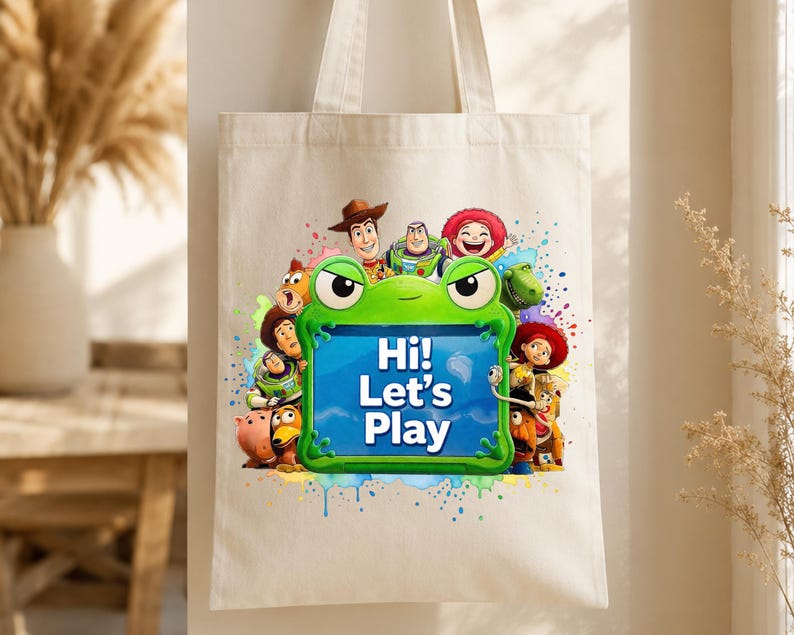 May include: A cream-colored canvas tote bag featuring a colorful graphic of Toy Story characters surrounding a bright green frog-shaped frame. The center of the frame contains the blue text "Hi! Let's Play" against a light blue background.