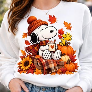 May include: A white crewneck sweatshirt featuring a graphic of a white dog wearing an orange knit hat and a plaid scarf, holding a mug with a heart. The design includes a yellow bird, orange pumpkins, a sunflower, and falling autumn leaves.