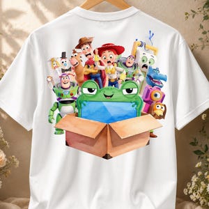 May include: A white t-shirt featuring a colorful graphic print of various Toy Story characters, including Woody, Jessie, and Buzz Lightyear, gathered around a brown cardboard box with a green frog-shaped object inside the box.