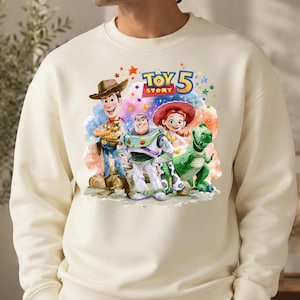 May include: A cream-colored crewneck sweatshirt featuring a colorful graphic print of Woody, Buzz Lightyear, Jessie, and Rex from Toy Story. Above the characters, the text reads Toy Story 5 in blue and yellow letters, surrounded by small stars.