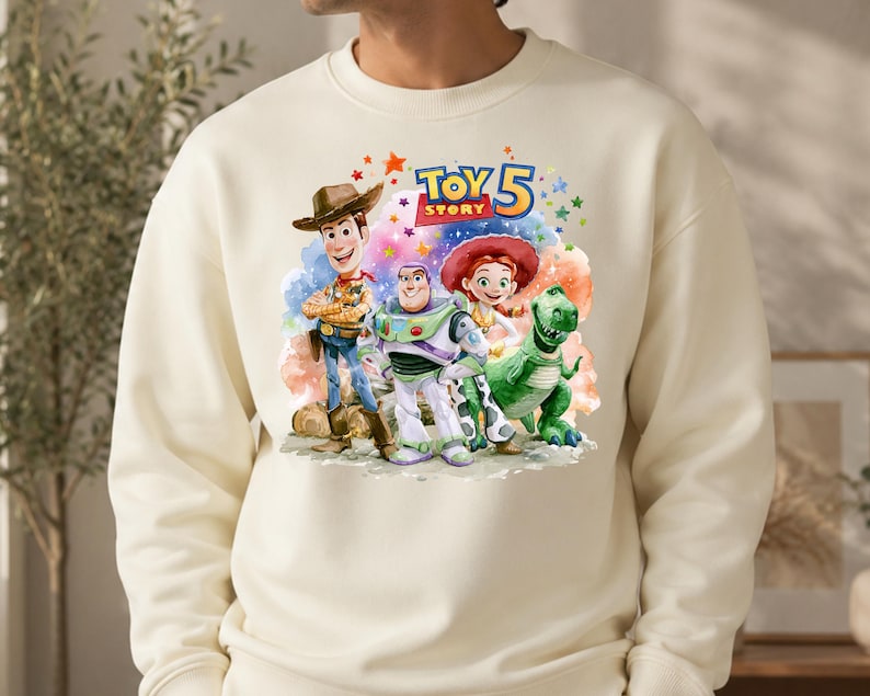 May include: A cream-colored crewneck sweatshirt featuring a colorful graphic print of Woody, Buzz Lightyear, Jessie, and Rex from Toy Story. Above the characters, the text reads Toy Story 5 in blue and yellow letters, surrounded by small stars.
