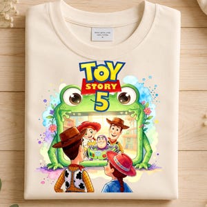 May include: A cream-colored t-shirt featuring a graphic print of a green frog-shaped frame containing Toy Story characters Woody, Jessie, and Buzz Lightyear. The text Toy Story 5 is printed in blue and red above the frame. The shirt has a tag.