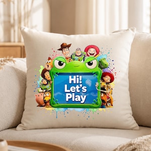 May include: A square off-white throw pillow featuring a central green frog-shaped frame with the text Hi! Let's Play in white. The frame is surrounded by a colorful graphic illustration of various Toy Story characters including Woody, Buzz Lightyear, and Jessie against a vibrant paint splatter background.