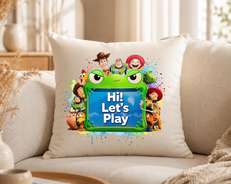 May include: A square off-white throw pillow featuring a central green frog-shaped frame with the text Hi! Let's Play in white. The frame is surrounded by a colorful graphic illustration of various Toy Story characters including Woody, Buzz Lightyear, and Jessie against a vibrant paint splatter background.