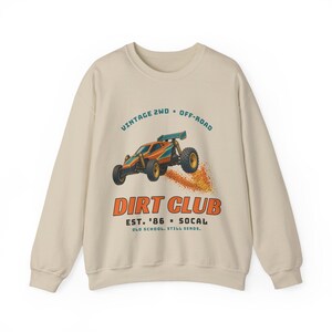 Puede incluir: Una sudadera beige de cuello redondo con un gráfico de un coche todoterreno a control remoto naranja y azul levantando polvo. El texto dice VINTAGE 2WD OFF-ROAD, DIRT CLUB, EST. '86 SOCAL y OLD SCHOOL. STILL SENDS. en azul y naranja.
