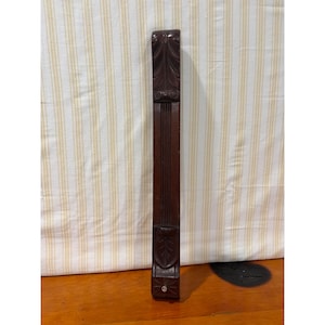 May include: A vertical, dark brown wooden furniture leg or architectural corbel featuring carved leaf motifs at the top and bottom, with a central section of vertical fluting and a small metal screw visible at the base of the piece.