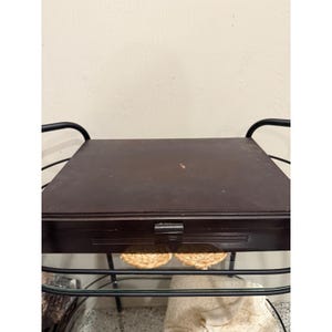 Può includere: Una scatola rettangolare in legno marrone scuro con coperchio piatto e una piccola chiusura centrale, caratterizzata da dettagli a scanalature orizzontali sul pannello frontale. La scatola è posizionata su una struttura in metallo nero.