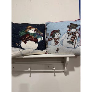May include: Two square throw pillows featuring woven winter scenes. The left pillow has a dark blue background with a snowman wearing a brown hat and scarf holding gifts. The right pillow has a light blue background with two snowmen.