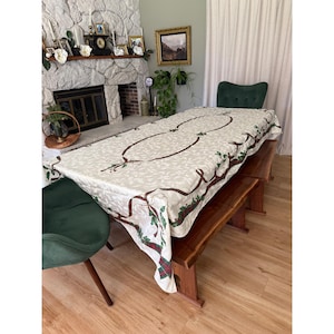 May include: A rectangular tablecloth featuring an off-white fabric with a subtle tonal leaf pattern. The design includes a central brown vine motif accented with green holly leaves and red berries, finished with a plaid ribbon border detail.