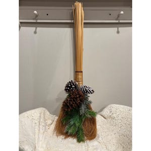 May include: A decorative broom featuring a long, light brown handle made of thin, straight wooden reeds. The base is adorned with a cluster of brown pinecones, some tipped with white, and green artificial pine needles tied together.