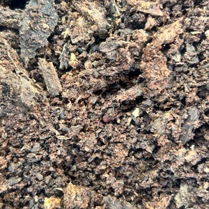 May include: A close-up view of dark brown organic potting soil featuring a coarse texture with visible wood chips, small bark fragments, and fine soil particles. The composition is dense and earthy, showing a mix of dark brown and black tones.