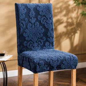 May include: A navy blue chair cover featuring a raised, textured damask pattern. The fabric is fitted over a high-backed chair with light-coloured wooden legs, providing a uniform appearance across the seat and backrest with a consistent floral motif.