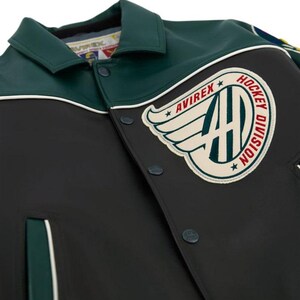 May include: A black and dark green jacket featuring a large circular patch on the chest with the text AVIREX HOCKEY DIVISION and a winged logo. The jacket has a pointed collar, snap button closures, and white piping accents along the shoulder seams.
