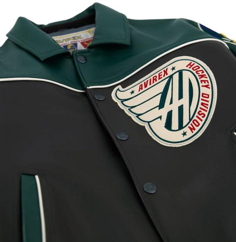 May include: A black and dark green jacket featuring a large circular patch on the chest with the text AVIREX HOCKEY DIVISION and a winged logo. The jacket has a pointed collar, snap button closures, and white piping accents along the shoulder seams.