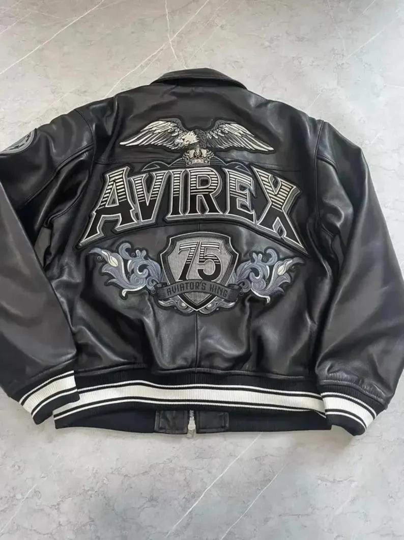 Op de afbeelding: Een zwart leren jack met een grote geborduurde adelaar, de tekst AVIREX en een schildvormig embleem met het getal 75 en de tekst AVIATOR'S KING op de achterkant. Het jack heeft een geribbelde kraag en boorden met witte en zwarte strepen.