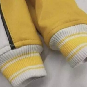 May include: A jacket sleeve featuring a yellow fabric body with a white panel and a thin black vertical stripe. The cuff is made of ribbed knit material in white with two horizontal yellow stripes, providing a snug fit at the wrist area.