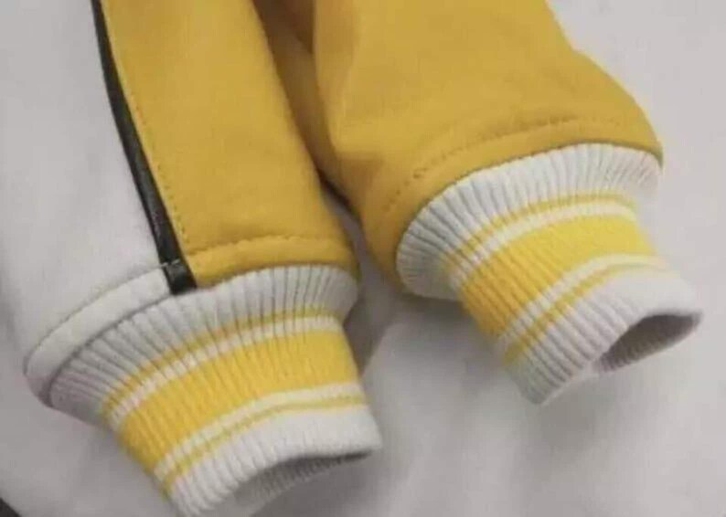 May include: A jacket sleeve featuring a yellow fabric body with a white panel and a thin black vertical stripe. The cuff is made of ribbed knit material in white with two horizontal yellow stripes, providing a snug fit at the wrist area.