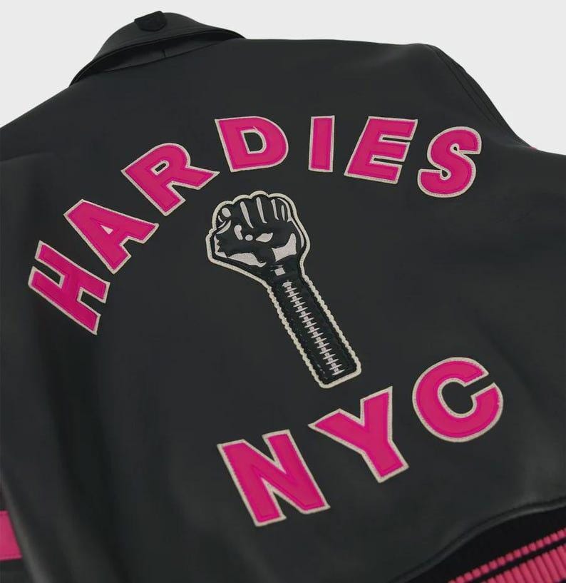 Peut inclure: Une veste noire avec le texte HARDIES en lettres roses arqu&eacute;es au-dessus d'un graphique repr&eacute;sentant un poing ferm&eacute; avec un motif de fermeture &eacute;clair sur l'avant-bras, et les lettres NYC en rose sous le poing, brod&eacute;s au dos du v&ecirc;tement.