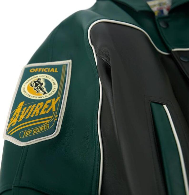 May include: A dark green and black leather jacket featuring white piping accents. A rectangular patch on the sleeve displays the text OFFICIAL, CANADIAN HOCKEY TEAM, AVIREX, and TOP SCORER, along with a graphic of a hockey player in a circular frame.