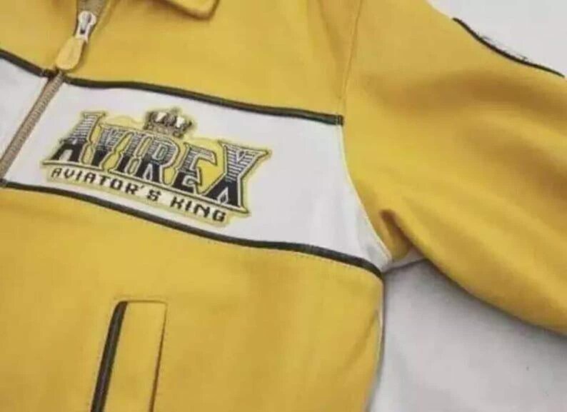 May include: A yellow jacket with a white horizontal chest stripe and a black zipper closure. The chest features an embroidered logo that reads AVIREX AVIATOR'S KING with a crown graphic. The jacket includes a collar and a vertical pocket.