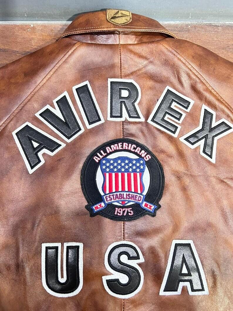 以下が含まれることがあります： 上部に大きな黒と白の文字でAVIREX、下部に同じ文字でUSAと記された茶色のレザージャケット。中央の円形パッチには、星とストライプが描かれた盾と、ALL AMERICANS ESTABLISHED N.Y. 1975という文字が表示されています。