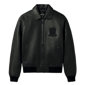 Op de afbeelding: Een zwart leren jack met een klassieke kraag, een volledige ritssluiting aan de voorzijde en geribbelde boorden bij de mouwen en zoom. Op de borst bevindt zich een ton-sur-ton geborduurde patch met de tekst AVIREX. Het jack heeft twee verticale zijzakken.