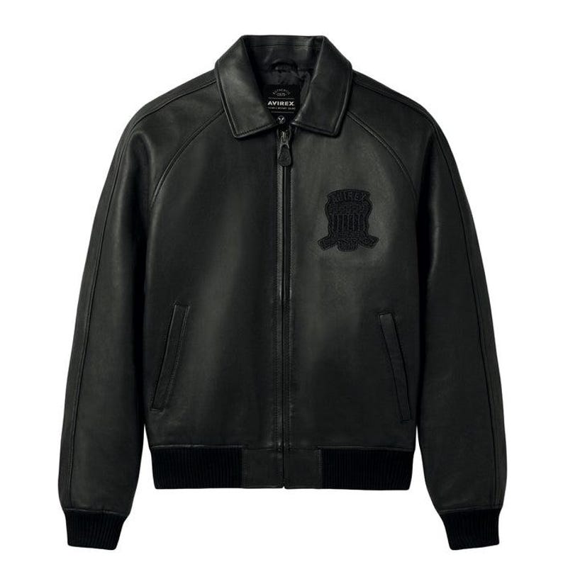 Op de afbeelding: Een zwart leren jack met een klassieke kraag, een volledige ritssluiting aan de voorzijde en geribbelde boorden bij de mouwen en zoom. Op de borst bevindt zich een ton-sur-ton geborduurde patch met de tekst AVIREX. Het jack heeft twee verticale zijzakken.