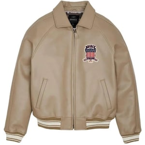May include: A beige bomber jacket with a full-length front zipper, a pointed collar, and ribbed cuffs and hem featuring white stripes. The chest displays an embroidered patch with the text AVIREX above a shield featuring the American flag.