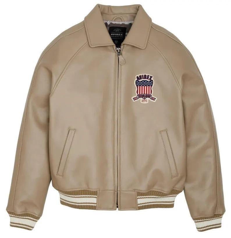 May include: A beige bomber jacket with a full-length front zipper, a pointed collar, and ribbed cuffs and hem featuring white stripes. The chest displays an embroidered patch with the text AVIREX above a shield featuring the American flag.