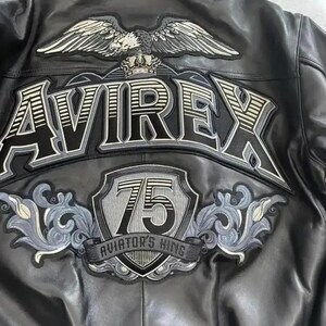 Op de afbeelding: Een zwart leren jack met een grote geborduurde patch op de achterkant. Het ontwerp bevat een adelaar, een kroon, de tekst AVIREX, een schild met het getal 75 en de tekst AVIATOR'S KING, geaccentueerd met grijs en wit decoratief krulwerk.