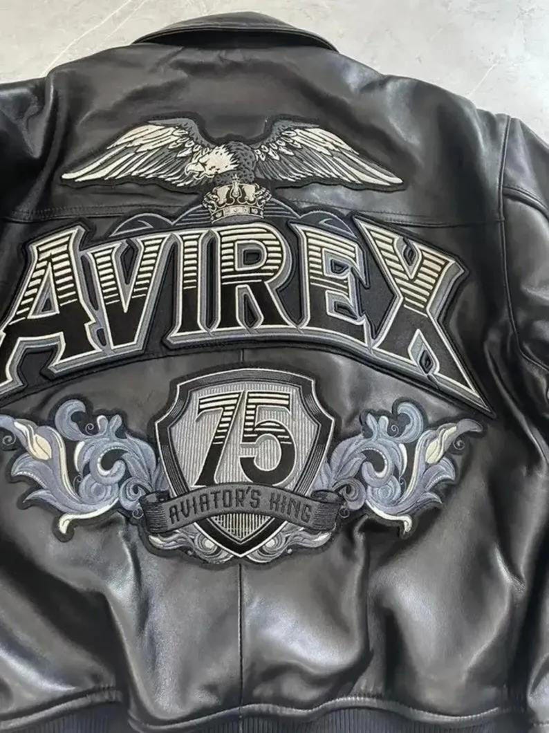 Op de afbeelding: Een zwart leren jack met een grote geborduurde patch op de achterkant. Het ontwerp bevat een adelaar, een kroon, de tekst AVIREX, een schild met het getal 75 en de tekst AVIATOR'S KING, geaccentueerd met grijs en wit decoratief krulwerk.