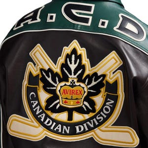 May include: A dark brown leather jacket featuring a green leather upper back panel with the text A.C.D. embroidered in black and white. A large embroidered patch displays a black maple leaf, a gold crown, and the text AVIREX CANADIAN DIVISION.