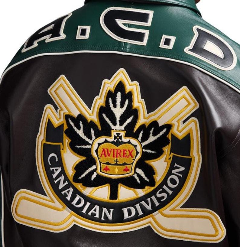 May include: A dark brown leather jacket featuring a green leather upper back panel with the text A.C.D. embroidered in black and white. A large embroidered patch displays a black maple leaf, a gold crown, and the text AVIREX CANADIAN DIVISION.