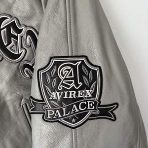 Puede incluir: Una chaqueta de cuero gris claro con un parche bordado en blanco y negro en la manga. El parche muestra un dise&ntilde;o de escudo con la letra A, hojas de laurel y el texto AVIREX PALACE sobre una cinta con una peque&ntilde;a estrella.