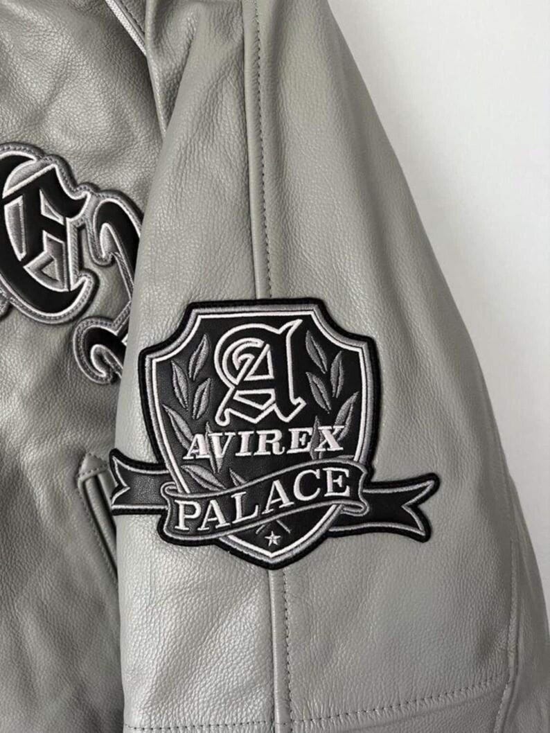 Puede incluir: Una chaqueta de cuero gris claro con un parche bordado en blanco y negro en la manga. El parche muestra un dise&ntilde;o de escudo con la letra A, hojas de laurel y el texto AVIREX PALACE sobre una cinta con una peque&ntilde;a estrella.