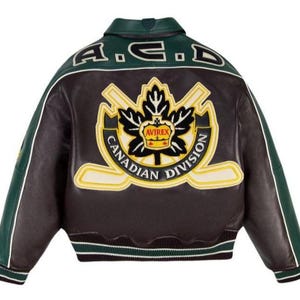 May include: A dark brown and green leather varsity jacket featuring a large embroidered patch on the back. The patch displays a black maple leaf, two crossed hockey sticks, a yellow crown with the text AVIREX, and CANADIAN DIVISION text.