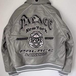 Puede incluir: Una chaqueta bomber de cuero gris con un gran texto bordado en la espalda que dice Palace New York, London, Tokyo y Avirex 75. La chaqueta incluye un dobladillo y pu&ntilde;os acanalados a rayas grises y blancas, con ribetes blancos en las costuras.