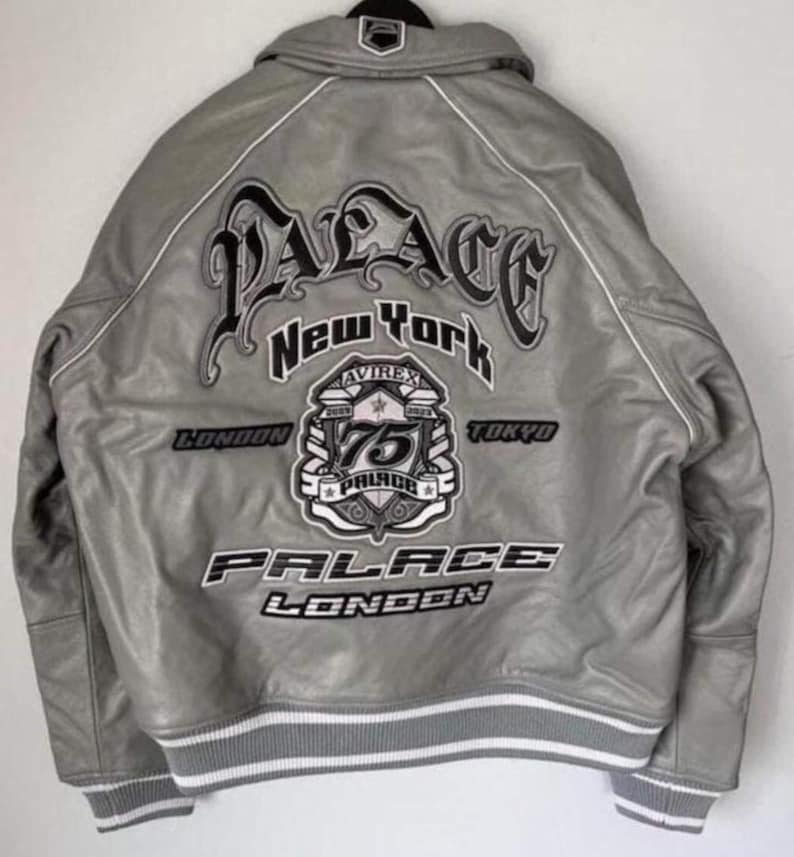 Puede incluir: Una chaqueta bomber de cuero gris con un gran texto bordado en la espalda que dice Palace New York, London, Tokyo y Avirex 75. La chaqueta incluye un dobladillo y pu&ntilde;os acanalados a rayas grises y blancas, con ribetes blancos en las costuras.