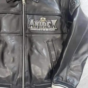 Op de afbeelding: Een zwart leren jack met een zilveren ritssluiting aan de voorzijde, een kraag en geribbelde boorden met witte strepen. Op de borst staat een geborduurde patch met de tekst AVIREX AVIATOR'S KING en een kroon, op de mouw een ronde 75-patch.