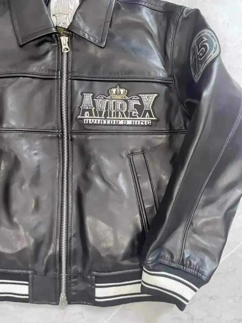 Op de afbeelding: Een zwart leren jack met een zilveren ritssluiting aan de voorzijde, een kraag en geribbelde boorden met witte strepen. Op de borst staat een geborduurde patch met de tekst AVIREX AVIATOR'S KING en een kroon, op de mouw een ronde 75-patch.