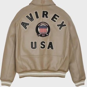 May include: A tan leather bomber jacket featuring a fold-down collar with a small logo patch. The back displays large black applique lettering reading AVIREX and USA, with a central circular patch showing the American flag and text 1975.